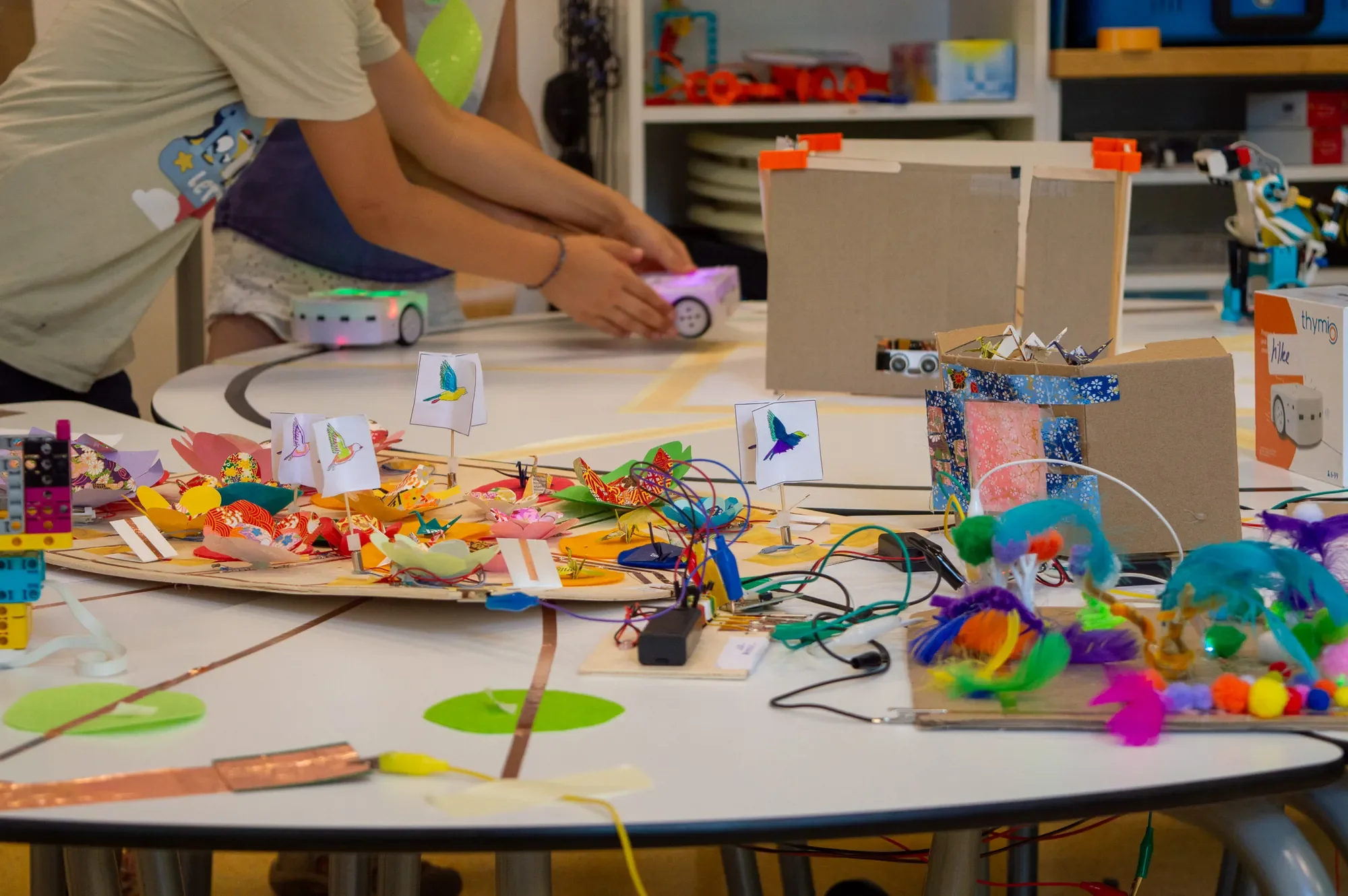 Papierblumenwiese mit Elektronik; dahinter Kinder mit Thymio-Robotern
